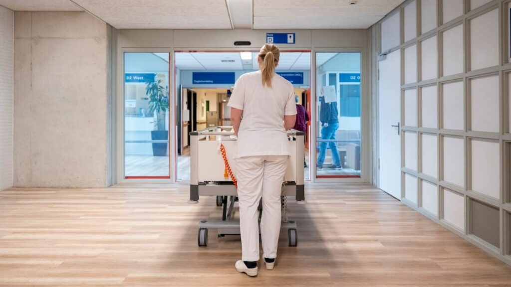 Foto van de achterkant van een vrouw die door het ziekenhuis loopt met een ziekenhuis bed.