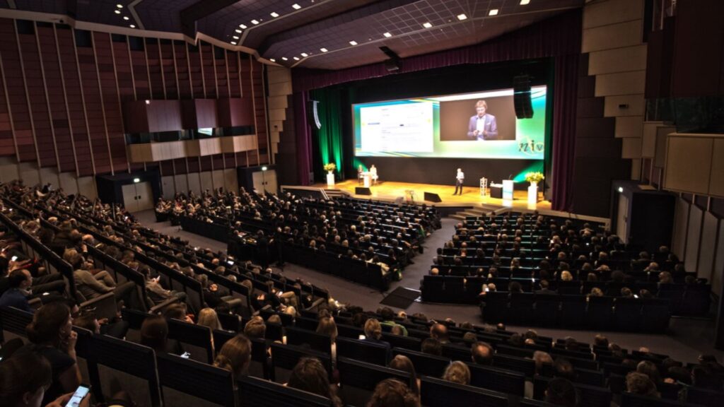 Foto van een volle zaal met mensen die kijken naar een presentatie van NIV